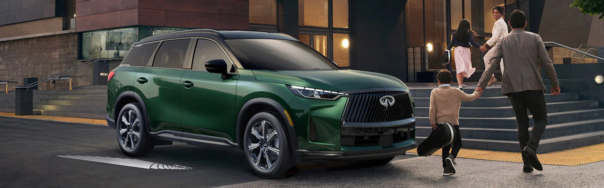Green Infiniti QX60 SUV parked outside a modern building with a family walking nearby.