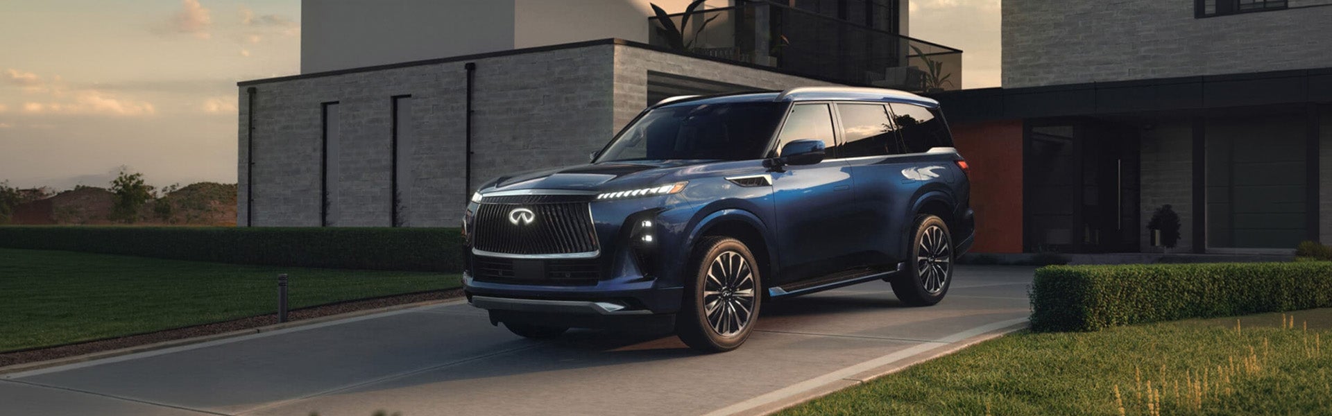 A dark blue Infiniti SUV parked in the driveway of a modern luxury home at sunset, featuring a large front grille, sleek LED headlights, and polished alloy wheels.
